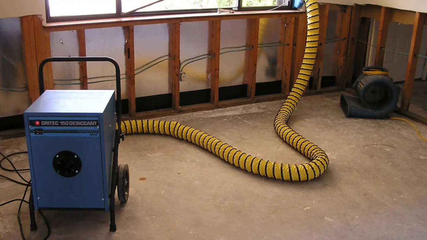 Dehumidifier with a yellow duct hose drying an opened wall cavity beside the baseboard
